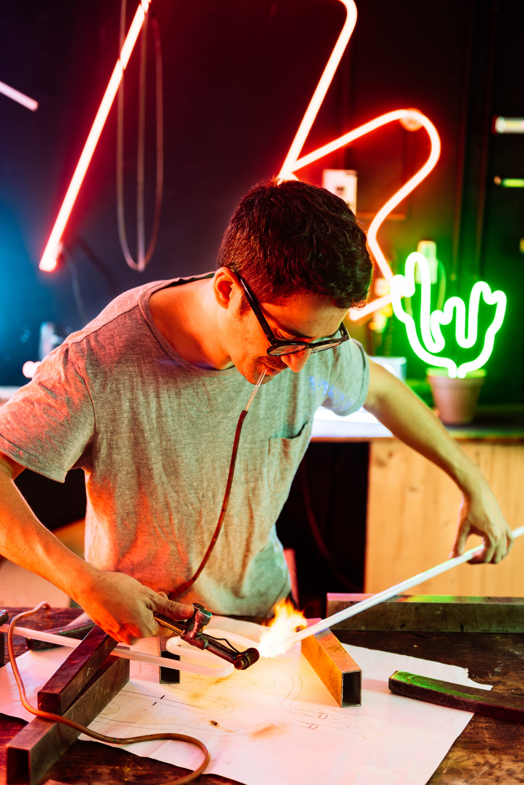 A man wearing glasses bends a neon tube with a torch, creating a custom neon sign in a workshop.